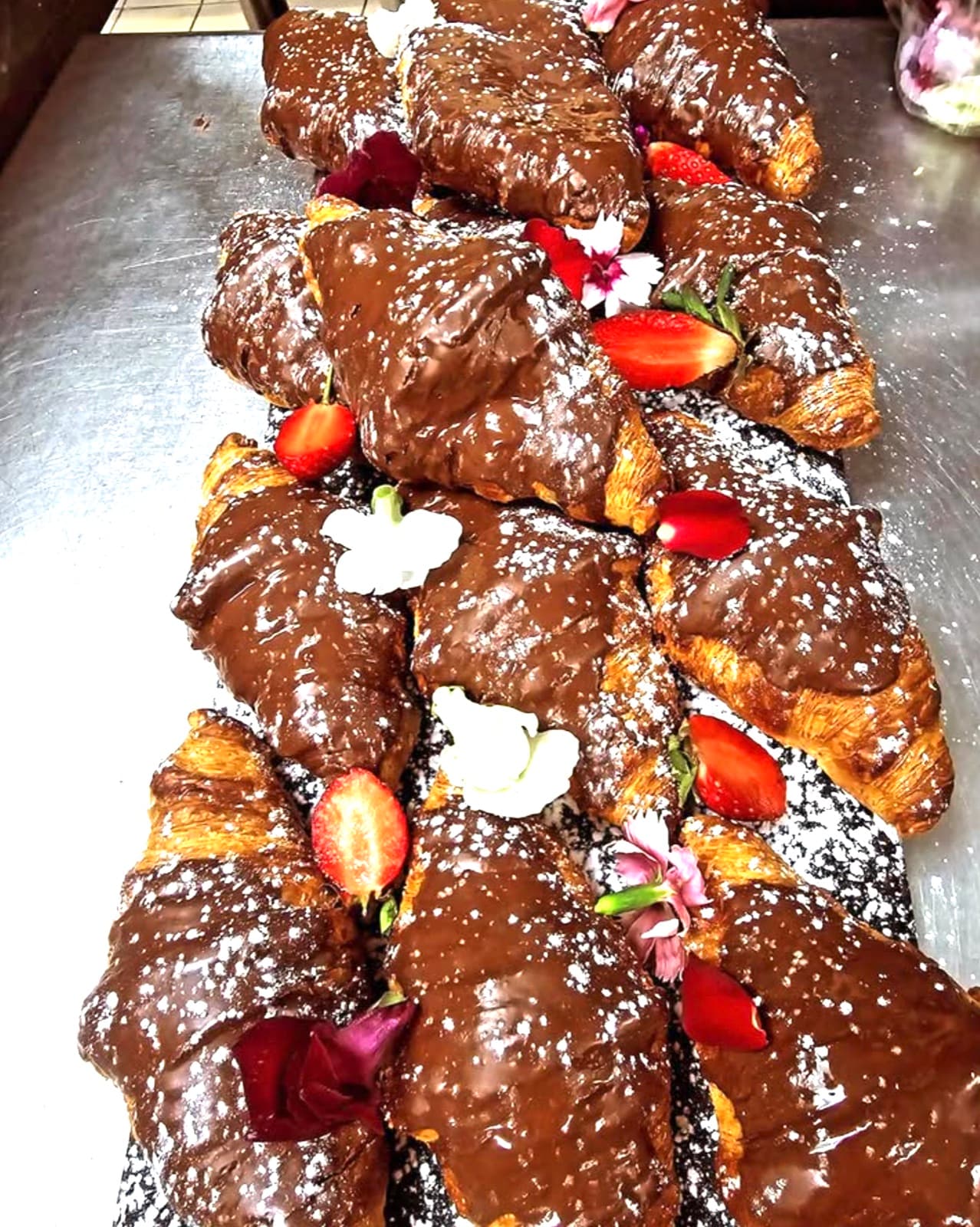 A row of chocolate-glazed croissants dusted with icing sugar, finished with strawberries and edible flowers.