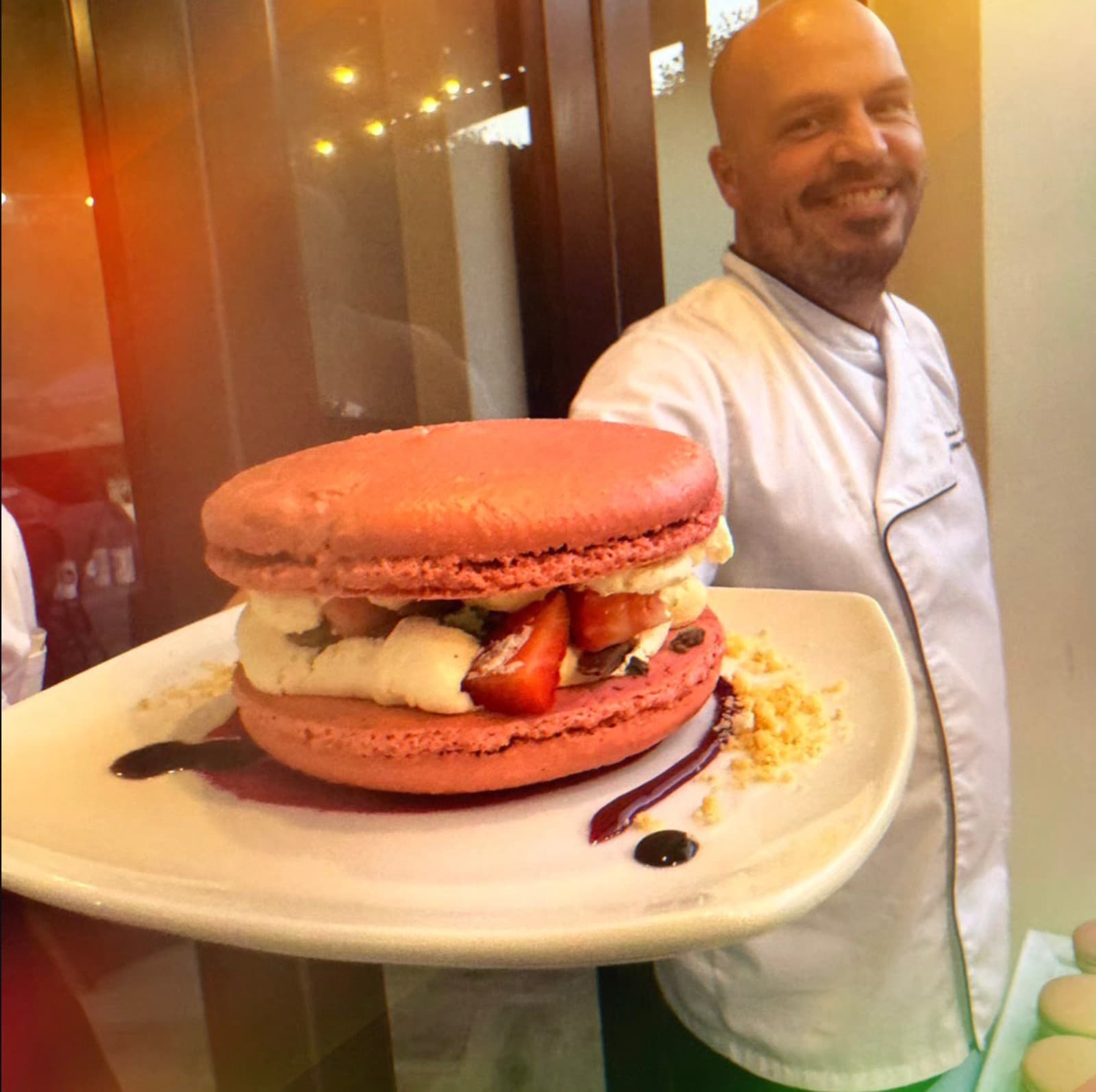 Chef Tristan smiling, presenting a giant strawberry-and-cream macaron on a plate.